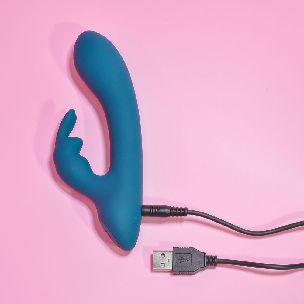 Top side view of the packaging and teal Playboy Pleasure Lil Rabbit vibrator, with the charger displayed on a pink background | Flirty Bay adult store and lingerie store